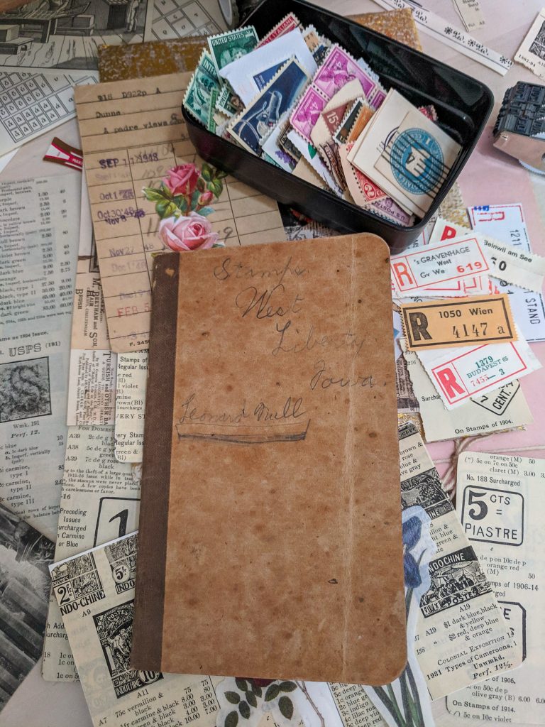 The cover of an old stamp album is displayed, lying on top of a messy desk of collage papers.